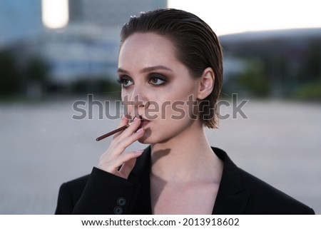 Attractive and elegant Caucasian young woman in a suit smoking a cigarette, unhealthy, outdoors on a summer day.