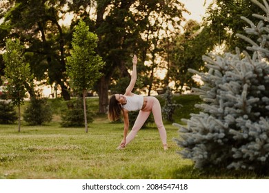 attractive, elegant woman do yoga and stands in Trikonasana pose while maintaining balance on green grass of lawn at sunset, concept of inner harmony. 