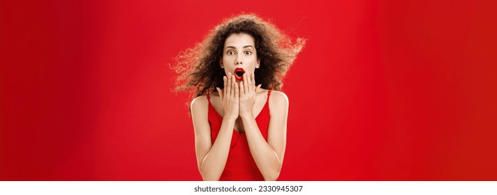 Attractive elegant young woman in red evening dress. with curly hair flicking on wind saying wow being impressed and thrilled covering opened mouth from amazement standing over red background.