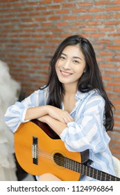 Attractive Emotional Smiling Asian Thai Woman Singing And Playing Acoustic Guitar. Beautiful Woman Singer Playing Musical Instrument Indoors. People, Music, Joy And Fun , She Looks At Camera