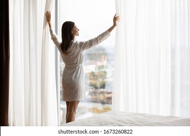 Attractive energetic happy young brunette woman wearing nightgown bathrobe, opening curtains on big panoramic window. Smiling lady enjoying starting of sunny day holiday vacation morning time.