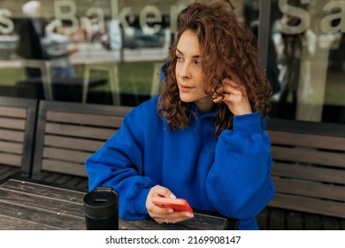 Attractive european brunette with curls holding phone in hands, smiling and looking aside while touching chin on blurred street background. Girl teenager uses smartphone for fun. - Shutterstock ID 2169908147