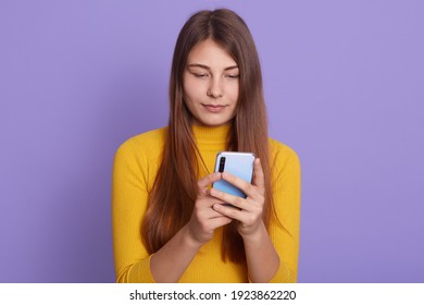 Attractive European girl wearing yellow shirt, holding smart phone and looking at its display, typing messages, posing isolated over lilac wall, using wireless Internet, sharing useful information.