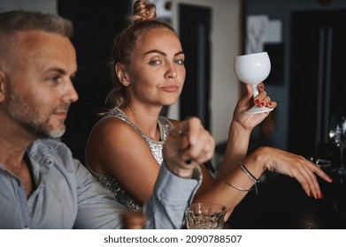 Attractive European Woman With A Drinking Glass In Hands, Looks At Camera Sitting At The Bar Close To A Handsome Caucasian Middle Aged Man, Talking, Having Fun Together, Flirting, Drinking Alcohol