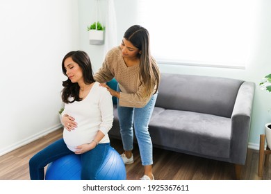 Attractive expectant mother sitting on a fitness ball in the living room and relaxing with a back massage from her lovely midwife