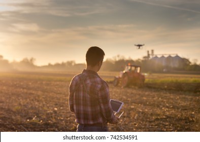 Attractive farmer navigating drone above farmland with silos and tractor in background. High technology innovations for increasing productivity in agriculture