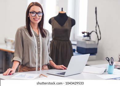 Attractive Fashion Designer Is Looking At Camera And Smiling While Working With Sketches And A Laptop In Her Office