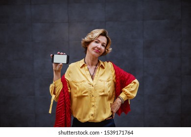 Attractive Fashionable Caucasian Apathetic Senior Woman Holding Smart Phone While Standing With Hand On Hip.