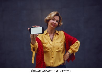 Attractive Fashionable Caucasian Apathetic Senior Woman Holding Smart Phone While Standing With Hand On Hip. Selective Focus On Hand With Smart Phone.