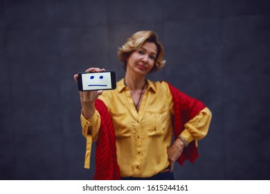 Attractive Fashionable Caucasian Apathetic Senior Woman Holding Smart Phone With Neutral Face On It While Standing With Hand On Hip. Selective Focus On Hand With Smart Phone.