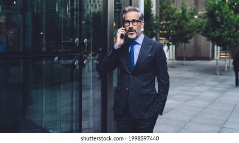 Attractive fashionable gray businessman with beard in stylish dark business suit talking on phone near office building in city street in New York