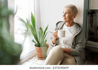 Attractive fashionable senior caucasian blond woman enjoying cup of hot herbal tea admiring beautiful view of noisy big city looking through window sitting on windowsill in in hoodie - Powered by Shutterstock