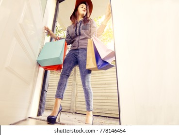 attractive fashionable woman with many shopping bags wearing jeans, a hat and high heels entering through the door. concept of consumerism and fashion shopping addiction. low angle full body view.