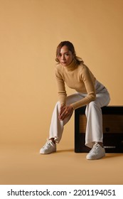 Attractive Fashionable Woman Wearing Trendy Sweater Sitting On A Vintage Radio, Studio Shot On Beige Background