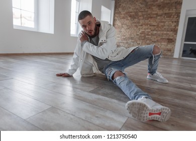 Attractive Fashionable Young Model Man With A Beard In A Stylish Jacket And Fashion Ripped Jeans With Sneakers Sits On The Wooden Floor In The Studio