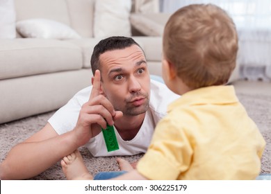 Attractive Father Is Lying On Flooring Near His Small Son. He Is Raising His Finger Up And Explaining Something To His Child With Seriousness. The Boy Is Listening To Him Attentively