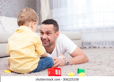 Attractive Father Is Playing With His Son And Smiling. He Is Lying On Flooring And Looking At The Boy With Love. The Toddler Is Sitting With Joy. Copy Space In Right Side