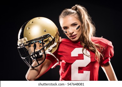 Attractive female american football player in uniform posing with helmet and looking at camera isolated on black
