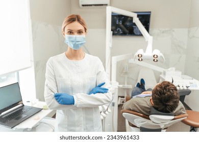 Attractive female dentist wearing protective mask, standing in modern dental office, arms crossed, looking at camera. Male patient sitting in dental chair. Health care, treatment concept - Powered by Shutterstock