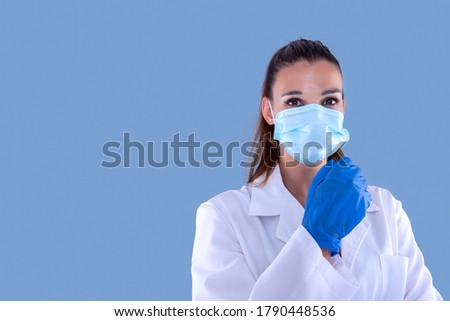 Attractive female doctor with virus protection mask and latex gloves looking straight ahead and blue background.