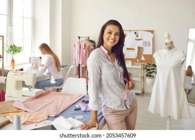 Attractive female fashion designer looking at camera and smiling while standing in her office. Woman with measuring tape around her neck posing standing in her own sewing workshop.