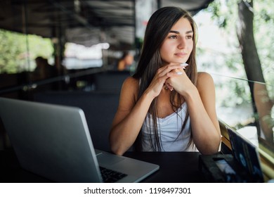 Attractive female freelance sitting at table front open computer in modern coffee shop. Young creative woman work on laptop while having breakfast on terrace, flare sun