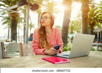 Attractive female freelancer hold smart phone while sitting at wooden table front open computer in modern coffee shop,young creative woman work on laptop while having breakfast on terrace, flare sun