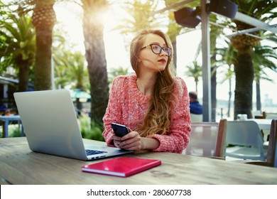 Attractive female freelancer hold smart phone while sitting at wooden table front open computer in modern coffee shop,young creative woman work on laptop while having breakfast on terrace, flare sun - Powered by Shutterstock