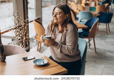 Mujer atractiva en gafas con una sonrisa tierno tomando un café mientras se relaja en un descanso. Mujer positiva en uso casual sonriendo y descansando con una taza de bebida caliente en un acogedor café. Concepto de desayuno sencillo