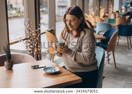 Attractive female in glasses with cute smile having a coffee while relax in in a break. Positive woman in casual wear smiling and resting with cup of hot drink in cozy cafe. Concept of easy breakfast