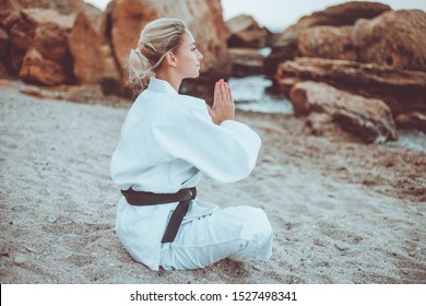 Attractive female martial artist in white kimono with black belt sits on sand and meditates on a wild beach - Powered by Shutterstock