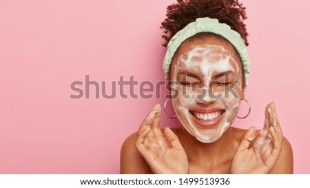 Attractive female model with crisp combed hair, closes eyes and gestures actively, feels pleased, washes face with cleansing foam, wears headband, wants to have well cared complexion and skin