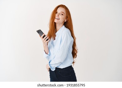 Attractive Female Model With Long Ginger Hair Listening Music On Mobile Phone With Wireless Earphones, Standing Happy Against White Background