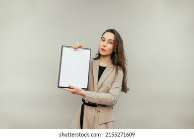 Attractive Female Office Worker Stands On A Beige Background With A Tablet In Her Hands With A White Sheet Of Paper And Shows To The Camera With A Serious Face.