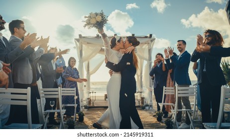 Attractive Female Queer Couple Walking Up The Aisle At Outdoors Wedding Ceremony Near Ocean. Two Lesbian Women In Love Share Happiness With Diverse Multiethnic Friends. Cute LGBTQ Relationship Goals.