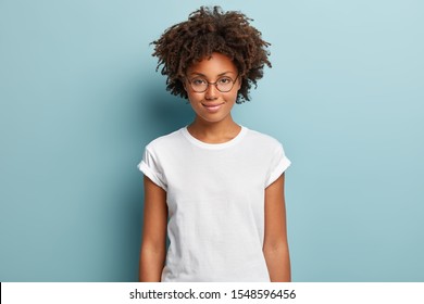 Attractive female student with curly hair, wears transparent glasses, white t shirt, stands against blue background, has calm face expression, tender smile, listens interlocutor with pleasure