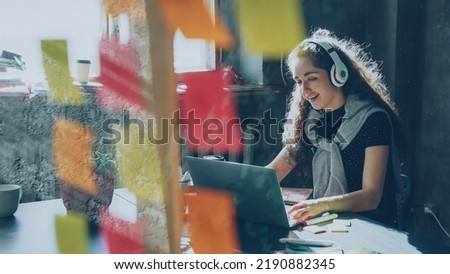 Attractive female student is listening to music with headphones dancing and singing while working with laptop computer indoors. Glassboard with bright colored stickers in foreground.