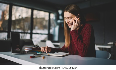Attraktive Studentin in der Universitätsbibliothek für das Schreiben von Hausaufgaben, die über Mobiltelefon mit Freunden kommunizieren mit Anwendung, modernen Laptop-Computer und Lehrbuch auf dem Tisch bleiben