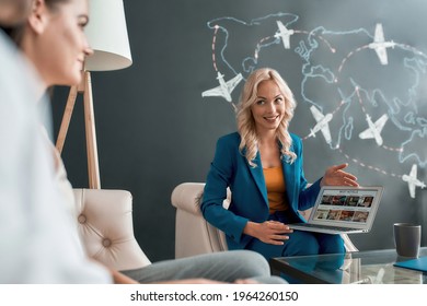 Attractive female travel agent looking at her clients, communicating with them in office and showing tours, using laptop. Tourism, travelling, business concept