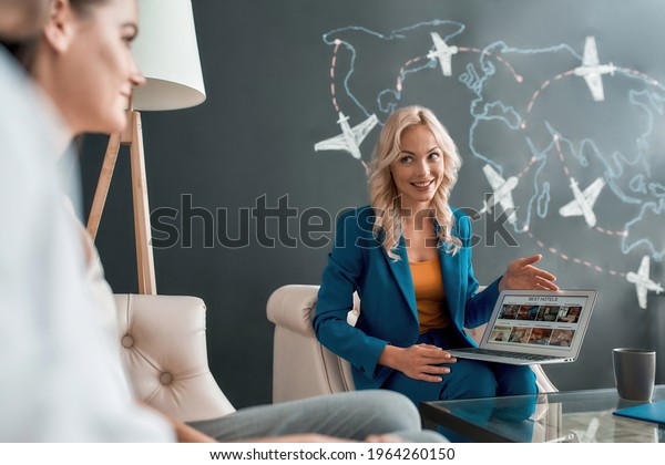 Attractive female travel agent looking\
at her clients, communicating with them in office and showing\
tours, using laptop. Tourism, travelling, business\
concept