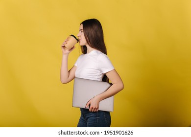 Attractive Female In Withe T-shirt Hold Closed Laptop And Drink Coffee. Side View. Place For Text. Woman Standing Isolated Over Yellow Background