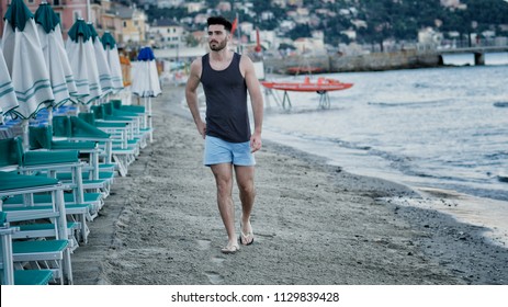Attractive fit athletic young man walking along the beach among lounge chairs and sun umbrellas, wearing black tank-top