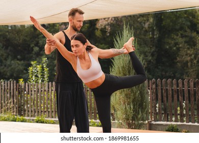 Attractive Fit Male Trainer Helps His Student With Stretching In Yoga Outdoors