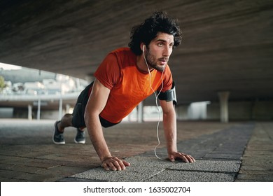 Attractive fit man listening to music in earphone doing push-ups exercises during workout outdoors - man showing perseverance and determination