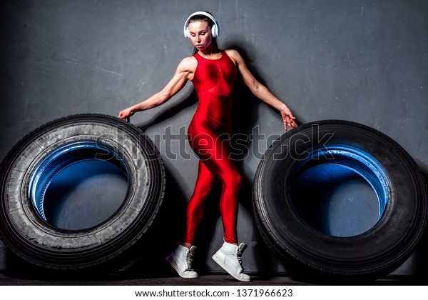 Attractive fit woman athlete in red sport costume\
and wireless headphones posing with a huge tires in the gym. Fit\
woman with big tire.