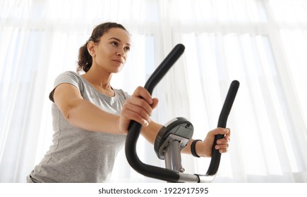 Attractive Fit Woman Exercising On A Spin Bike At Home On A Beautiful Sunny Day. Cardio Workout. Fitness At Home. Close-up