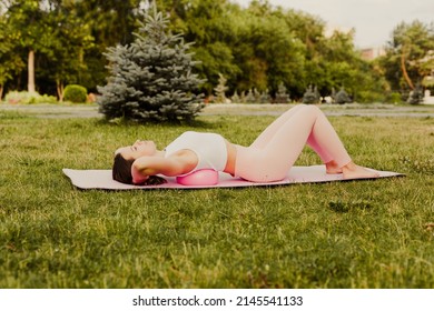 attractive, flexible Caucasian woman do pilates with ball and stands in bridge for stretching spine on green grass of lawn at sunset.