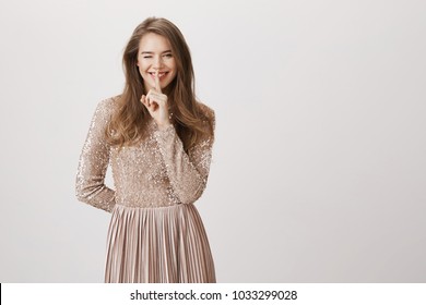 Attractive Flirting Caucasian Woman In Stylish Evening Dress Making Shhh Gesture And Hiding Something Behind Back, Winking And Smiling Broadly, Expressing Excitement Over Gray Background.