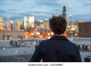 Attractive, Forty Something Man, With Long, Curly Brunette Hair, On A Rooftop, In The City, With Cityscape In The Distance. Dusk And Sunset Lights. Staring Into The Distance. Contemplating The Future.