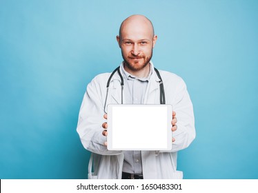 Attractive Friendly Bald Bearded Smiling Doctor Holds Tablet With White Screen Looking At Camera Isolated On The Blue Background Mockup